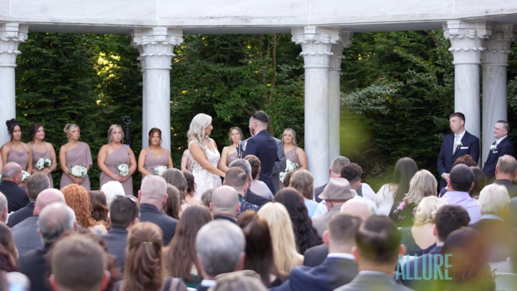 Stephen and Lindsey stand hand in hand, exchanging vows in front of towering marble pillars that encircle them. The atmosphere is filled with happiness as numerous guests and bridesmaids look on with joyful anticipation.